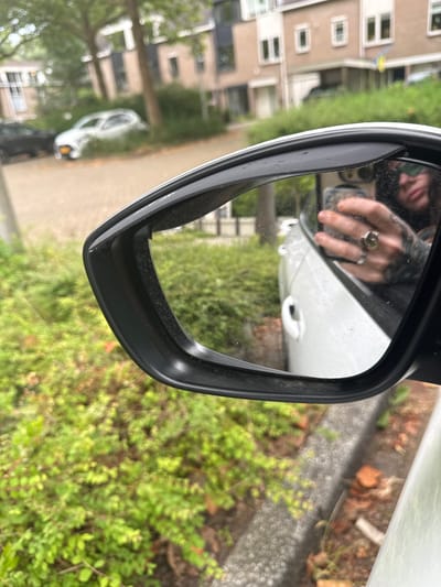 The image captures a close-up view of a car's side mirror, reflecting a person's hand holding a smartphone. The person's hand is adorned with at least two rings, one being a prominent silver ring with a unique design. The reflection also shows a glimpse of the person's face and tattoos on their arm. The car appears to be white and is parked outdoors.
The background, visible through the mirror and in the surrounding environment, suggests a residential area. We can see houses with multiple windows and light-colored facades, typical of modern European architecture. Greenery, including bushes and trees, is abundant, indicating a well-maintained outdoor space. The presence of a paved area, possibly a driveway or sidewalk, and some scattered bricks or debris on the ground suggest a typical urban or suburban setting.
While the exact location in Amstelveen, Netherlands, cannot be definitively determined from the image alone, the architectural style of the houses and the overall landscaping are consistent with residential neighborhoods found in the Netherlands. The image is taken during daylight, with overcast or diffused lighting conditions. The car's side mirror is slightly dirty, with visible smudges and water spots on its surface.