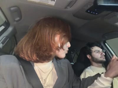 Two people are sitting in the front seats of a vehicle. On the left side of the image is a man with dark hair and a beard, wearing a white shirt under a black jacket. On the right side is a person with red hair, wearing a light-colored t-shirt and a grey jacket. The interior of the vehicle includes a dark grey dashboard with buttons and a warning label. The location cannot be determined based on this image.