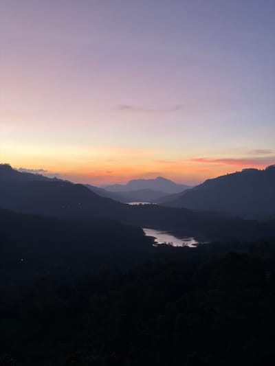 The image depicts a panoramic landscape view during twilight in Ramboda, Sri Lanka. The sky transitions from a deep purple at the zenith, through lavender and pink hues, to a vibrant orange and yellow gradient along the horizon. Several layers of silhouetted mountains dominate the scene, receding into the distance and appearing progressively lighter due to atmospheric haze. In the middle ground, two reflective bodies of water, likely lakes or reservoirs, are visible, mirroring the lighter tones of the sky. The immediate foreground and lower mountain slopes are covered in dense, dark vegetation, appearing almost black. Sparse artificial lights, suggesting distant human habitation, are faintly visible on the slopes of the mountains in the mid-left sector. No discernible people, specific objects, or actions are evident within the frame.