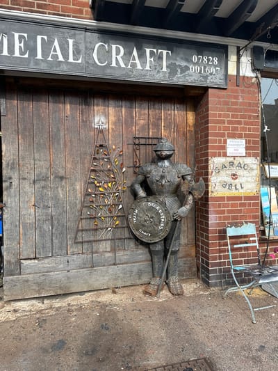The image displays the exterior of a "Metal Craft" shop in Broadstairs, Thanet, United Kingdom. A life-sized dark metallic knight statue, bearing the name "REGGIE" on its chest, stands prominently. The knight holds a circular shield in its left hand, embossed with "BROADSTAIRS METAL CRAFT" and a fleur-de-lis, and an axe resting on the ground in its right hand. Behind the statue are weathered, vertically paneled wooden double doors serving as the main entrance. Above these doors, a dark signboard displays "METAL CRAFT" in white letters and phone numbers "07828 001607". To the immediate left of the knight, a metal garden trellis decorated with numerous small, multi-colored metal leaves is affixed to the wooden doors. To the right of the wooden doors, a section of red brick wall is visible. Attached to this wall are a white "PRIVATE PROPERTY NO PARKING" sign and a faded "GARAGE SELL" sign. A light blue, distressed metal folding chair is positioned against the brick wall on the far right. The ground is a dark, uneven paved surface, with a metal grate partially visible in the bottom right corner.