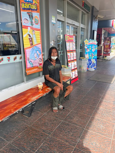 A person wearing a black t-shirt, dark green pants, and flip-flops is sitting on a low wooden ledge in front of a 7-Eleven store in Ban Ko Pru, Thailand. The person has long dark hair and is wearing a white face mask. They are resting their arms on their legs and appear to be looking towards the viewer. Next to the person is an open cup, possibly containing food or drink.

The store has a large orange banner across the storefront with various cartoon characters and text in Thai, including "ELEVEN" and "ALL ลาดาว แลกพรีเมียม". There are also posters visible inside the store, one featuring a roasted chicken with the price "129-". The ground is tiled with brown and black patterned tiles, and there is sunlight casting shadows. The scene appears to be set during the daytime. The overall mood is quiet and everyday, with the person resting outside the convenience store.