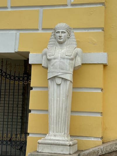 A horizontal, eye-level shot shows a white marble statue of a pharaoh mounted on the outside wall of a light-yellow building. The statue has a human face and head with a striped headdress, and its arms are crossed over its chest. The statue is wrapped in a white marble sheath that is striped vertically. The statue sits on a white base with vertical and horizontal striations. The building behind the statue is light yellow with vertical white columns. A section of black and gold fencing is visible beneath the statue. The building is located in the city of Arkhangel'skoe, Russia.