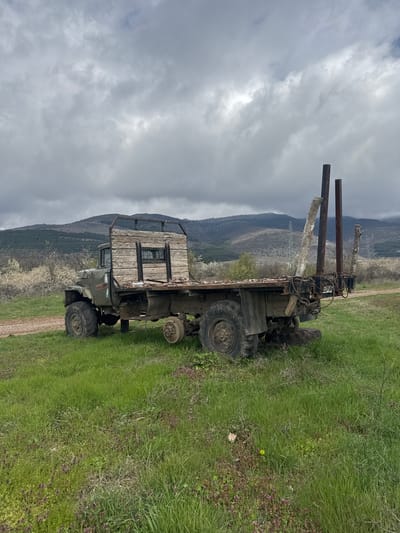 The image depicts an old, dilapidated logging truck parked on a grassy field in Gorna Grashtitsa, Bulgaria. The truck's cab is made of weathered wood with a visible window. It has multiple large, worn tires. Extending from the front of the truck are two sets of poles, one rusty metal, and the other made of tree limbs. A mountain range is visible in the background under a cloudy sky. The field is overgrown with grass and small wildflowers. A dirt path is visible on the right side of the image. There are no people visible.