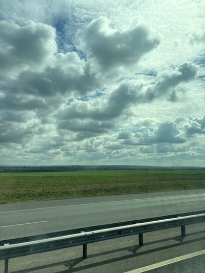 The image depicts a vast, open landscape under a dramatic, cloud-filled sky. In the foreground, a grey asphalt road runs horizontally across the frame, bordered by a metal guardrail with vertical supports casting shadows on the road. The road has a white dashed line marking its center. Beyond the road, a wide expanse of green fields stretches towards a distant, tree-lined horizon. The sky is filled with a complex arrangement of white and grey clouds, with patches of blue sky visible in between. The overall impression is one of a rural or semi-rural setting, possibly viewed from a moving vehicle. There are no people or specific city landmarks visible in the image that would indicate it is located in Menzelinsk, Russia.