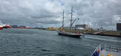 In the city of Den Helder, Netherlands, a tall ship with two masts and a black hull is sailing on a waterway. The ship has numerous lines and rigging, and a Dutch flag is flown from the main mast. The deck of the ship is populated with several people. A small boat is tethered to the stern of the tall ship, leaving a wake in the water. Several other vessels are moored in the background, including a large red ship and a white ship with a red stripe. Industrial buildings and structures are visible along the waterfront, with a cloudy sky overhead. In the foreground, the edge of another boat, painted blue and gray with a white railing and orange life preservers, is visible.