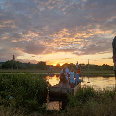 In Căzănești, Moldova, a boat carrying five children navigates a tranquil body of water during a sunset. The sky above is a dramatic canvas of orange and pink clouds, illuminated by the setting sun. The children, appearing to be of various ages, are positioned within the boat, some standing and others leaning over the edge. The boat itself is rustic, possibly made of wood, and rests low in the water. Surrounding the waterway is lush green vegetation, including tall grasses and reeds that line the banks. In the background, a treeline with rolling hills forms the horizon, and a distant church steeple with a blue dome is visible. A utility pole with wires stands in the middle ground, suggesting the proximity of human settlement. The overall scene depicts a peaceful, natural setting at dusk with the activity of children enjoying the water.
