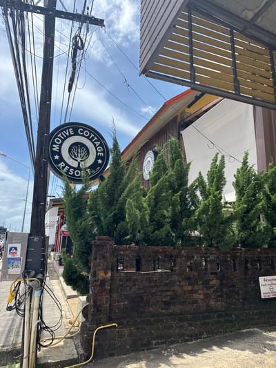 The image shows a street scene in Ban Bang Niang, Thailand. A black circular sign with white lettering reads "MOTIVE COTTAGE RESORT CO.,LTD" and features a drawing of a tree. The sign is attached to a wooden utility pole covered in numerous black electrical wires. Several tall, conical green trees stand behind the sign, partially obscuring a brick wall and a building with a yellow roof and wooden siding. A white banner is visible on the building. To the left of the utility pole is a grey electrical box with stickers, including one with a QR code and a phone number. The street is paved with concrete, and a yellow hose lies on the ground. In the background, a white sign with red text and a circular no-trespassing symbol is attached to the brick wall. The sky is blue with scattered clouds.