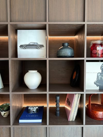 This image, captured in Marche-lez-Écaussinnes, Belgium, depicts a section of a contemporary wooden shelving unit constructed with a grid-like pattern of individual cubbies. Some shelves are equipped with horizontal LED strip lighting embedded along their back edges.

From left to right and top to bottom, the visible contents of the shelves include:

*   **Top Row:** A print featuring a side profile of a dark, vintage Buick Grand National coupe on a light background; a dark, textured, squat ceramic vase; and a clear glass jar filled with red, spherical items, bearing a handwritten label that reads "BLUE TOP 2021."
*   **Middle Row:** A matte white, spherical ceramic vase; a black vinyl record with an orange label, standing vertically and leaning against the rear of the cubby; and a print displaying a monochrome image of a person with the text "sade love deluxe."
*   **Bottom Row:** Two stacked, glazed ceramic bowls with green and brown patterns; a blue-covered book titled "Ocean" with a silver-toned vintage rangefinder camera resting horizontally on its cover; a dark, sculpted object composed of stacked, irregular spheres; and a stack of vinyl records, with the top album cover displaying "IGOR" in pink lettering, adjacent to a partially visible red woven basket.

No individuals or active interactions are present in the scene.