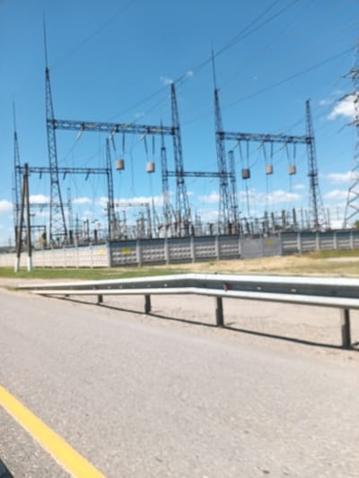 This media file depicts an outdoor scene of a power substation under a clear blue sky with scattered white clouds. The primary subjects are the large metal structures of the power substation, including towering pylons, crossbars, and insulators. Cylindrical components, possibly transformers or arresters, hang from the crossbars. The substation is enclosed by a concrete fence, with some lighter colored structures visible behind it.
In the foreground, a paved road with a yellow line marking the edge runs horizontally across the frame. A metal guardrail with black posts is positioned between the road and the substation. The overall impression is of an industrial infrastructure facility.
The time of day appears to be daytime, given the bright sunlight and clear skies. The weather seems pleasant and sunny. There are no people visible in the frame.
No text is discernible in the image. The location context provided is Krymsk, Russia, suggesting this is a power substation in that region. The image is slightly blurred, which affects the clarity of fine details.