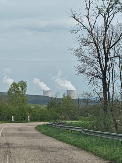 A rural road curves through a landscape with two nuclear cooling towers in the distance. Steam rises from the towers. Green hills and trees populate the mid-ground. A guardrail lines the road's edge. Bare trees are visible in the foreground on the right, and verdant trees are on the left. The sky is overcast. The location is in the vicinity of the Cattenom Nuclear Power Plant near Thionville, France.
