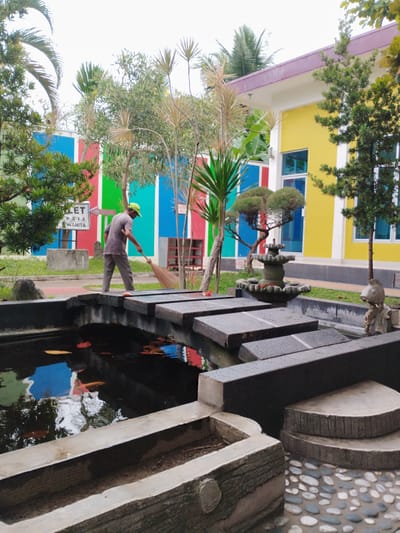 A man in a grey t-shirt and pants, wearing a green cap, sweeps a stone bridge over a koi pond. The pond contains several orange koi fish and fallen leaves. The bridge is made of dark grey rectangular stones, some of which are wet. In the background, a colorful building with yellow and purple accents is visible, featuring large blue windows. A striped fence in vibrant red, blue, and green colors runs along the building's perimeter. Tropical plants, including palm trees and a bonsai, are scattered around the scene. A small tiered fountain sits in the pond. To the left of the man, a sign reads "TOILET PRIA WANITA," indicating restrooms. The sky is overcast, suggesting a cloudy day. The overall setting appears to be a garden or park area, possibly part of a public facility or private residence. The activity depicted is routine maintenance.