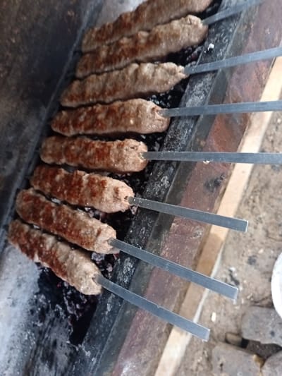 Several metal skewers filled with kebab meat are cooking on a grill. The kebabs are elongated and slightly browned. Black charcoal lies beneath the skewers. The grill itself is made of brick and metal. The background features blurry outdoor elements.
