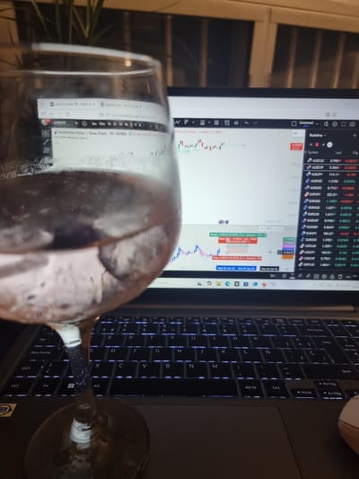 The image captures an indoor scene at night, illuminated primarily by the glow of a laptop screen. In the foreground, a wine glass, appearing partially filled and exhibiting condensation, occupies a significant portion of the lower-left to mid-right frame, partially obscuring the laptop screen.

The laptop is open, displaying what appears to be a financial trading or charting platform, likely TradingView, given the visible URL "www.tradingview.com/chart/". A candlestick chart for the currency pair AUDCHF is prominent, showing price movements. To the right of the chart, a watchlist displays various currency pairs including AUDCAD, AUDCHF, AUDJPY, AUDNZC, AUDUSD, CHFJPY, EURUSD, EURAUD, EURCAD, EURCHF, EURGBP, EURJPY, EURNZD, USDCAD, USDJPY, USDCHF, and GBPCAD, along with their last prices and percentage changes. Textual overlays on the chart indicate "Target: 0.00322 (0.569%) 12.2, Amount: 1250.78" and "Open: -0.00161, Qty: 77881". Dates like "Wed 10 Jun 26" and "Mon 29 Jun 26" are also visible on the chart timeline.

The laptop's backlit keyboard, featuring a Spanish layout with keys like "Ñ", "Blog Mayús", "Inicio", and "Alt Gr", is visible on the right side of the frame. A small portion of a white or wooden computer mouse can be seen near the top right corner of the keyboard. A blurred green plant is subtly visible in the background on the left. The scene suggests an individual is engaged in financial analysis or trading, possibly in the evening, accompanied by a drink.