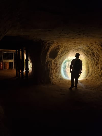 A person is silhouetted in a low tunnel, illuminated by a light source at the end of the tunnel. The walls are rough and textured. Lower in the image, a wooden structure with metal bars is visible. The image depicts a view inside the Historical Mine "Karlsbrunn" in Homburg, Germany.