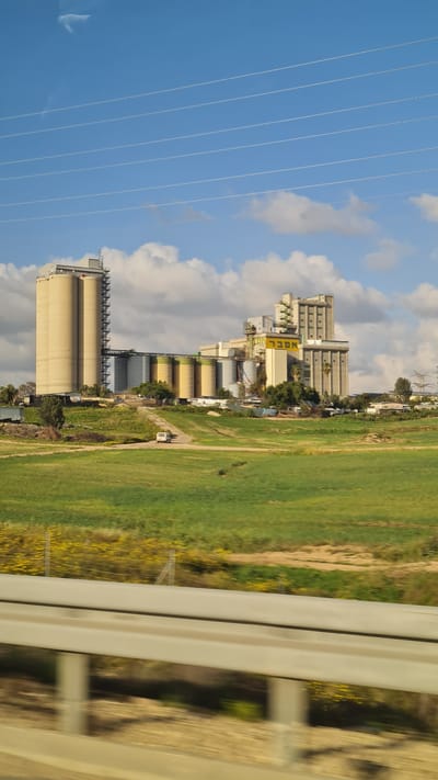 Captured from a moving vehicle, likely a train, the scene presents a landscape in Bnei Shimon Regional Council, Israel. The prominent features include an industrial complex with tall concrete silos, connected to buildings displaying the inscription "NOCL". To the right, a field with a mix of green and yellow vegetation extends, bordered by a narrow pathway. High-tension power lines stretch across a blue sky with scattered clouds on the left. The view is partially obscured by the vehicle's window frame on the right.