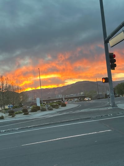 The image depicts an urban landscape during sunset in Reno, Nevada, United States of America. The upper sky is predominantly dark gray and blue-gray, gradually transitioning into vivid orange, red, and yellow hues near the horizon due to low sunlight illuminating clouds. A range of dark, rugged mountains is silhouetted against the brightly colored sky across the mid-ground.

In the foreground, a multi-lane asphalt road with white solid and dashed lane markings curves to the right. A gray metal pole stands on the right side, supporting a vertical traffic light assembly displaying a red signal. Below this, a pedestrian signal displays an illuminated red "Don't Walk" hand symbol. A blank rectangular sign is mounted above the traffic light.

Further into the scene, a concrete overpass or bridge spans horizontally, carrying traffic. At least three vehicles are discernible on the roads; one light-colored sedan with illuminated headlights is visible traveling away from the viewer on the road beneath the bridge. Multiple streetlights are active along the roads and on the bridge. A tall, gray streetlight pole stands prominently in the mid-ground, adjacent to a white rectangular utility box. Landscaped islands featuring green bushes, rocks, and some bare-branched trees are situated between road segments. Trees with sparse branches are visible on the far left. No individuals are clearly identifiable.