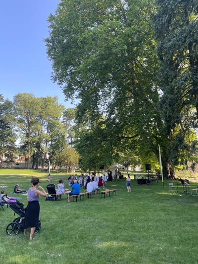 The image captures an outdoor event in Stupava, Slovakia, likely within the grounds of the Stupava castle park, given the presence of mature trees and manicured lawns characteristic of such a historical location. A group of people, some dressed in traditional folk costumes, are gathered on a grassy field. In the foreground, a woman with dark hair in a bun, wearing a purple tank top and a long black skirt, stands next to a large stroller. To her right, several other individuals are seated on wooden benches, facing a stage area in the background.
The stage area features a white tent, audio equipment including speakers, and musical instruments. More people, some in traditional attire with elaborate headwear and dresses, are arranged in front of the stage. A few individuals are standing and walking around the field, while others are seated on the grass or on blankets. The backdrop consists of lush green trees, providing shade, and a clear blue sky. The overall scene suggests a cultural performance or festival taking place on a sunny day.