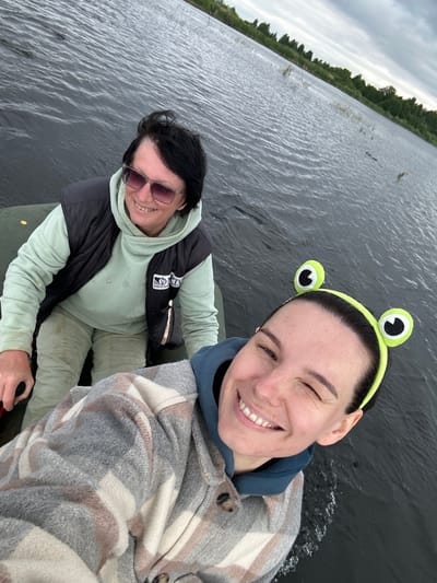 Two individuals are in a boat on a body of water, likely a lake or river, with a shoreline visible in the background. The person in the foreground is a woman with dark hair wearing a grey and white plaid jacket and a blue hooded sweatshirt. She is smiling and has a frog-themed headband on. The person behind her is an older woman with dark hair, wearing sunglasses, a black vest over a light green hooded sweatshirt, and tan pants. She is also smiling. The water appears choppy, and the sky is overcast. The location is identified as Dubrova, Russia.