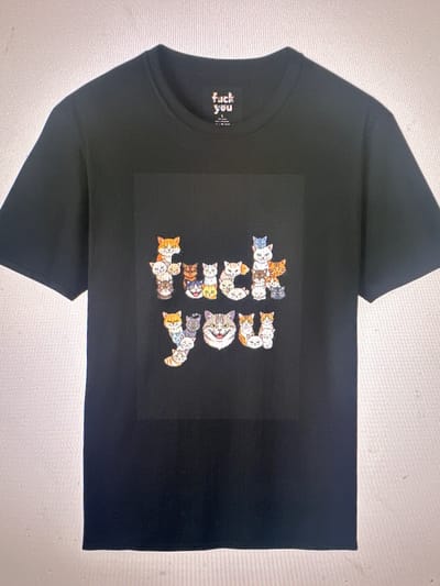 The image shows a black t-shirt with a graphic design printed on the front. The graphic is a word spelled out by a collection of cartoonish cats. The word "FUCK YOU" is formed by cats stacked in different positions. The t-shirt is lying flat on a surface. No specific location in Hallandale Beach, United States of America is identifiable in the image.