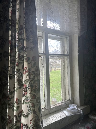 A view through an old, white-painted window with peeling paint. The window is partially covered by lace curtains at the top left and floral-patterned curtains at the bottom. Outside the window, green grass and trees are visible. A clear plastic container is affixed to the window frame at the top right. A glass bottle is seen in the window. The location is Студзёнкаўскі сельскі Савет, Belarus.