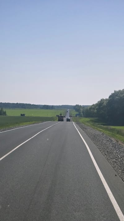 The image depicts a straight, asphalt road with white lane markings, leading towards a distant horizon. The sky is clear and pale blue. On either side of the road, there are green fields and trees. In the distance, several vehicles are visible on the road, including a red truck. The scene appears to be set in a rural area. There are no identifiable buildings or landmarks that would place this image specifically in the city of Актюбинский, Russia. The environment suggests a clear, sunny day.