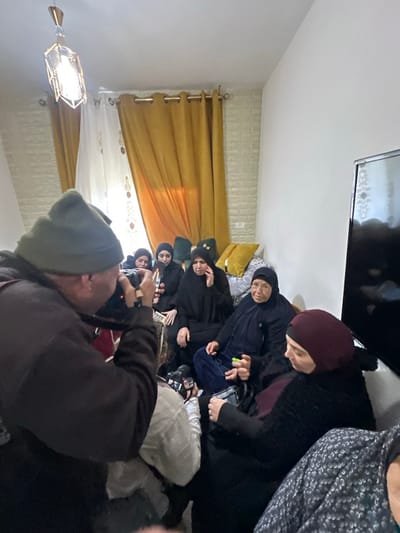 The image captures an indoor gathering in Dura, Palestinian Territory. In the foreground, a male individual, seen from behind, wearing a dark coat and a dark green knit cap, holds a camera device to his eye, appearing to film or photograph a group of women. Seven women are visible, seated on a light-colored sofa in the mid-ground and background. They are predominantly dressed in dark clothing and head coverings. One woman in black, central to the seated group, has her left hand raised to her face, indicating potential distress. An older woman with glasses, seated to her right, holds a small, bright green object in her right hand. Another woman to the left holds a smartphone vertically. The room features a textured light-colored brick-patterned wall on the left, partially covered by yellow and white curtains, and a plain white wall on the right with a mounted dark flat-screen television. A geometric pendant light fixture hangs from the ceiling.