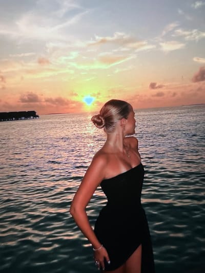 A young woman with blonde hair styled in a bun, wearing a black strapless dress, stands facing away from the camera, looking out at the ocean during sunset. The sun is low on the horizon, casting a warm orange and yellow glow across the sky and reflecting on the water. In the distance, there are overwater bungalows. The scene depicts a serene moment at a coastal resort. The image does not provide any information about the city of Montfermeil, France.
