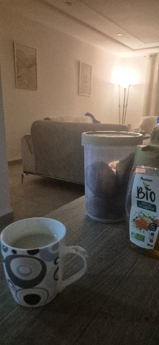 An indoor shot displays a mug with grey circle patterns lying on a wooden floor. Next to it sits a clear plastic container, partially filled with what appears to be dark brown food. Above the container is a bottle labeled "BIO," lying horizontally. A grey couch is visible in the background, near two rectangular wall art pieces on a white wall, illuminated by a light source.