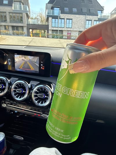 The scene is captured from inside a vehicle, specifically a Mercedes-Benz, showing its dark-toned dashboard and a portion of the passenger side. A human hand with manicured nails, featuring French tips and small embellishments, occupies the foreground, holding a green Red Bull "LIME GREEN EDITION" energy drink can. The can's label displays text including "Curuba-Fleur de sureau" and "Curuba-Vlierbloesem". On the dashboard, an infotainment screen displays a rear-view camera perspective with yellow guiding lines and a red boundary line. Below this, three circular air vents with chrome bezels exhibit blue internal ambient lighting. A control panel with illuminated red indicators is positioned beneath the vents. A red and silver Red Bull can is partially visible in a storage compartment to the lower left. Through the windshield, the exterior environment in Temse, Belgium, is visible. This includes two modern, multi-story buildings with light-colored facades, multiple windows, and glass balconies. The building on the right features a dark, pitched roof with skylights. An archway or entrance, guarded by yellow bollards, is situated between the two structures. Sparse, bare trees are present in the background. The sky is clear blue, indicating daytime conditions.