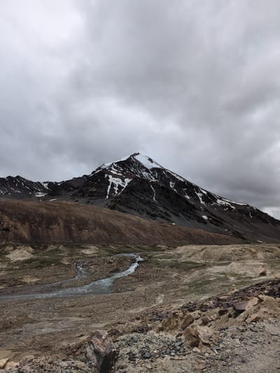 The image depicts a mountainous landscape under a cloudy sky. A prominent peak, partially covered in snow, dominates the background. The slopes of the mountains are rugged and rocky, with patches of brown earth and sparse vegetation. In the foreground, a winding river or stream flows through a rocky, arid terrain. The environment appears to be a high-altitude, remote area, characterized by its raw, natural beauty and the starkness of the geological formations. There are no people or specific structures visible in the scene. The location is identified as Killingsarai, India.
