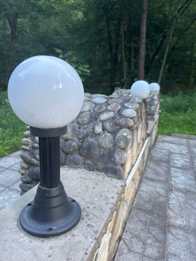 A close-up, eye-level outdoor shot captures a pathway in Lisnyky, Ukraine, featuring a stone wall and a series of globe lights. In the foreground, a black lamppost with a white globe light is mounted on a concrete surface. The lamppost base is circular with visible screw holes. The stone wall, constructed from various-sized, rounded river stones, extends into the background and is topped with two more white globe lights, one partially obscured. A light-colored trim runs along the top of the wall. To the right of the wall, a paved pathway with a geometric pattern leads away from the viewer, with a textured concrete surface. The background reveals a dense forest with trees and green foliage under a cloudy sky.