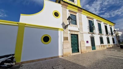 This is a high-angle, wide shot of the side of a white building with yellow trim, possibly the Faro Municipal Museum, located in Faro, Portugal. The building has a distinctive curved architectural element on the left side, with a yellow circular window. There are two more circular windows on the left wall. The main facade of the building features several green-paned windows and balconies with wrought-iron railings. A Portuguese flag hangs from one of the balconies. A large green door is visible in the center of the building. The ground in front of the building is paved with cobblestones. A white scooter is parked on the left side of the image. The sky is blue with scattered clouds. The overall scene suggests a sunny day in a historic European city.