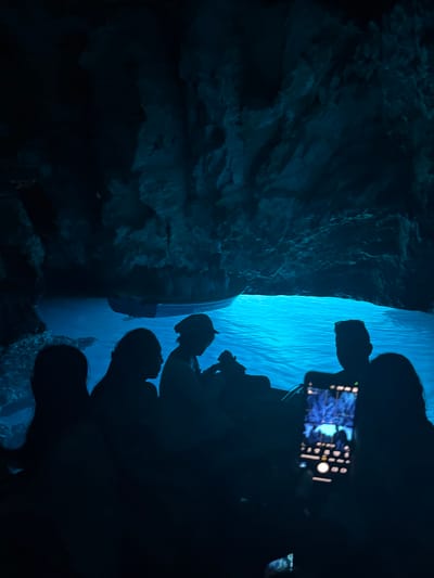 The image depicts the interior of the Blue Cave, a sea cave located off the coast of the island of Vis, near the town of Komiža, Croatia. The cave is illuminated by a surreal, vibrant blue light that reflects off the water, creating a magical atmosphere. Silhouetted figures of people are visible in the foreground, seated in a boat, their forms dark against the luminous blue water. One person in the foreground is holding up a smartphone, capturing the scene. The cave's rocky walls are visible in the background, dimly lit by the same ethereal blue glow. The overall impression is one of a captivating natural wonder, experienced by visitors in a boat.