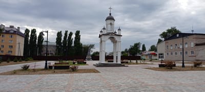 The image captures an outdoor scene in Kirsanov, Russia, featuring a central white archway structure topped with domes and a cross, resembling a religious monument or a decorative gazebo. The structure is situated in a paved public square with patterned brickwork. Benches are placed around the square, some with small shrubs and dry grass patches. In the background, there are several buildings, including a multi-story yellow apartment building on the left, a row of tall, slender trees, and a brick building on the right with multiple windows. The sky is overcast with gray clouds. The overall impression is of a public space, possibly a park or a town square, under a cloudy sky.