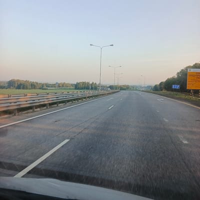 The image shows a highway in Uchkhoza Sel'khoztekhnikuma, Russia. The highway is paved and has white lane markings. There are streetlights on either side of the highway. In the distance, there are trees and fields. A yellow sign with Russian text is visible on the right side of the road. The sky is clear and blue. The image appears to have been taken from a moving vehicle.