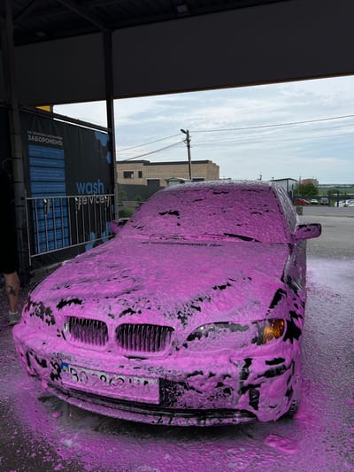At a self-service car wash bay in Velyki Hai, Ukraine, a dark-colored sedan, likely a BMW, is undergoing a wash. The vehicle is covered uniformly in a thick, bright pink cleaning foam. The front of the car, including its characteristic kidney grilles, headlights, and a partially obscured Ukrainian license plate (beginning "BO 2063 HX"), is visible. Pink foam is actively dripping from the vehicle onto the wet, dark ground surface.

To the immediate left of the car, the lower left leg and foot of an individual are visible, clad in a light gray sneaker. A dark tattoo, resembling an insect, is present on the individual's calf.

Behind the car, a dark blue informational banner displays Ukrainian text under the heading "НА ТЕРИТОРІЇ АВТОМИЙКИ ЗАБОРОНЕНО:" (On the territory of the car wash it is FORBIDDEN:), followed by a list of prohibited actions, including washing with the engine running or washing the engine itself. The words "wash service" are prominently displayed on the banner.

The car wash bay features an open metal frame structure supporting a light-colored ceiling. Beyond the bay, the sky is overcast with scattered clouds. In the background, various buildings are visible, including a light brown brick structure, a darker building, and other distant industrial or commercial properties. A utility pole with overhead power lines stands against the skyline. Several parked vehicles of varying colors are discernible in the distance.
