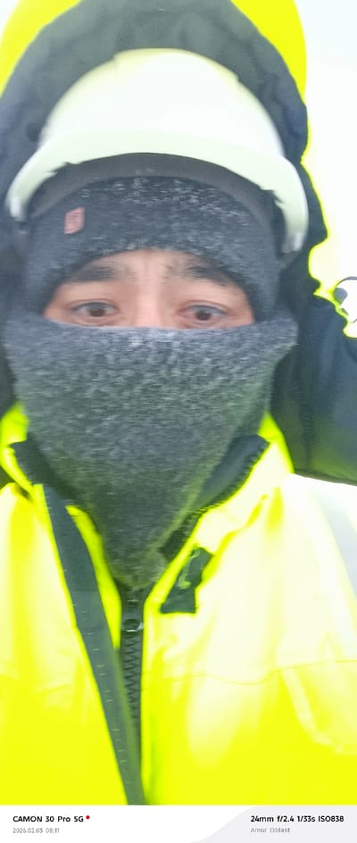 A close-up shot shows a person bundled up in winter gear. The individual wears a white hard hat, a gray knitted hat, and a gray scarf covering the lower part of their face, revealing only their eyes. They are also wearing a bright yellow jacket with a black zipper. The text overlay indicates that the photo was taken with a CAMON 30 Pro 5G, with specific camera settings and location information (Amur Oblast) noted. The date is 2026.02.03, at 08:11.