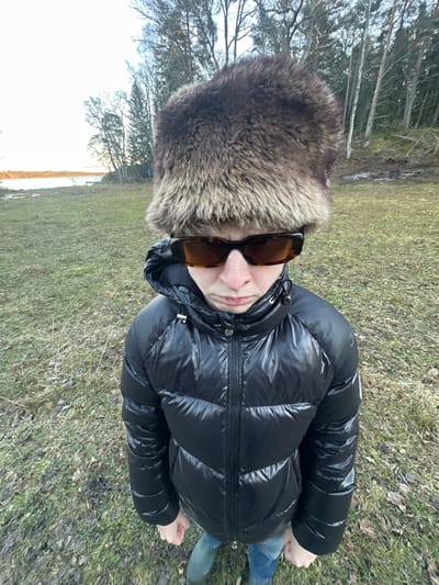 A single individual stands in an outdoor, natural environment within Boda, Sweden. The person, appearing young, is centered in the frame, facing the camera directly from a slightly elevated perspective. They wear a large, shaggy brown fur hat, rectangular tortoise-shell patterned sunglasses, a black shiny puffer-style winter jacket with a hood, blue denim jeans, and green rubber boots. The individual's mouth is visibly pursed in a slight frown, and both hands are clenched into fists at their sides. The ground consists of sparse, dry grass with scattered dead leaves. In the background to the left, a body of water is visible under a light, overcast sky, bordered by a treeline composed of mixed deciduous and coniferous trees. A small, dark patch of disturbed earth or ashes is present on the ground near the treeline on the right side of the image.