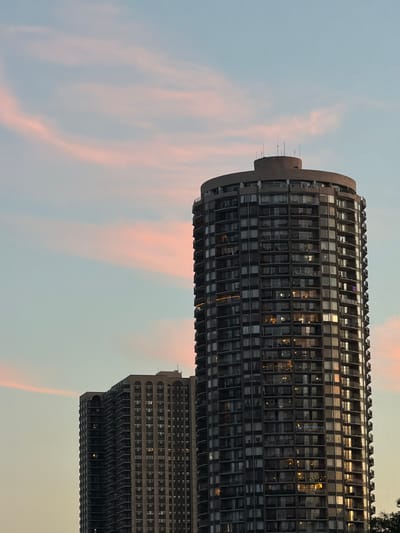A low-angle, full shot captures two tall residential buildings in Edgewater, USA, against a soft, pastel sky with wisps of pink clouds. The building on the right is a circular, modern high-rise with a dark facade and numerous balconies. Lights are visible in many of its windows, suggesting occupancy. The building on the left is rectangular, appearing older with a lighter-colored facade and also featuring balconies. Both buildings are silhouetted against the sky, with the primary focus on their architectural details and the fading light of dusk.