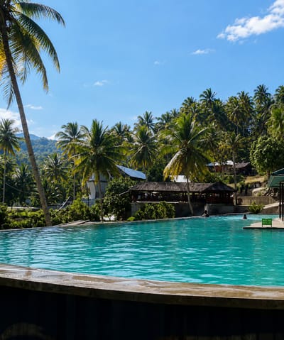 In Silaiyajae, Indonesia, a bright, clear day illuminates a luxurious infinity pool situated amidst lush tropical vegetation. The pool's turquoise water is calm, reflecting the vibrant blue sky dotted with wispy white clouds. Several palm trees, their fronds swaying gently, frame the scene, with one particularly large palm on the left casting a long shadow.
In the background, a cluster of buildings with thatched roofs and more modern structures are nestled among the trees, suggesting a resort or recreational area. Some of these buildings appear to be open-air structures, possibly restaurants or relaxation areas. A gentle slope of verdant hillside rises behind the buildings, further enhancing the tropical ambiance.
A few people are visible in the pool, their bodies partially submerged in the refreshing water, indicating a leisurely atmosphere. One person is swimming in the main pool, while another appears to be in a separate, shallower section marked with a sign that reads "KOLAM ANAK" (Children's Pool). The foreground of the image is dominated by the edge of the infinity pool, a concrete structure that creates a sleek, modern contrast with the natural surroundings. The overall impression is one of tranquility and relaxation in a beautiful, tropical setting.