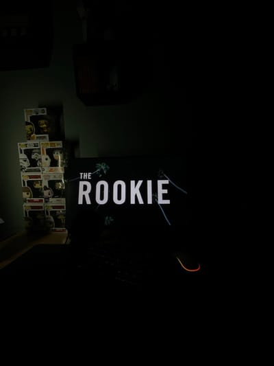 The image depicts a dimly lit indoor scene in Wolfisheim, France, primarily focused on a computer monitor and adjacent desk area. The monitor, positioned centrally, displays the title "THE ROOKIE" in white capital letters against a dark background, accompanied by stylized light blue palm tree graphics. Below the monitor, a dark keyboard is partially visible. To the right of the keyboard, a computer mouse with an illuminated orange-red outline rests on a dark surface. To the left of the monitor, a stack of at least six Funko Pop! collectible figures in their original boxes is visible on a surface, likely a desk or shelf. Specific figures include one with a golden appearance and several resembling Star Wars characters, such as a Stormtrooper. The background wall to the left of the figures is a dark, muted green or grey color. The overall environment is dark, with the monitor's display serving as the primary light source for the illuminated elements. No individuals are present in the frame.