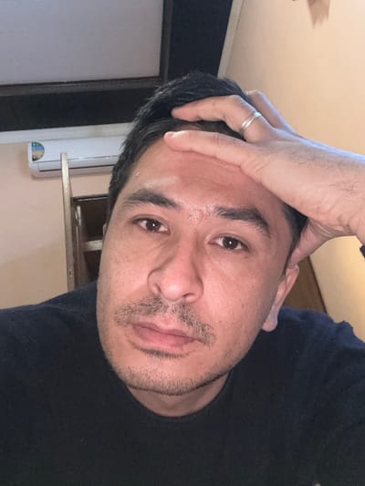 An indoor, close-up photograph depicts an adult male located in Baygundi, Uzbekistan. The subject has dark hair, brown eyes, and exhibits light stubble on his upper lip and chin, forming a mustache and beard. He is wearing a dark, possibly black, crew-neck garment and is looking directly at the camera with a neutral expression. His left hand is raised, with fingers resting against his forehead and scalp. A silver-colored ring is visible on the ring finger of his left hand. The background features a light-colored wall. In the upper left portion of the image, a white wall-mounted air conditioning unit with a blue and white label is visible. Above the air conditioning unit, a dark rectangular opening is present, and to its right, a vertical section of a light-colored wooden structure or frame is partially seen.
