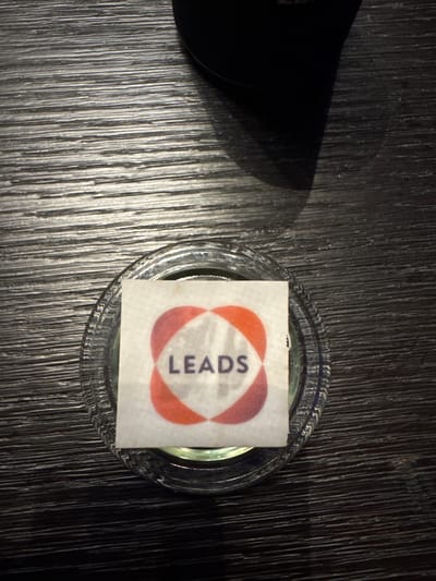 A square sticker with the word "LEADS" printed on it is placed on top of a clear glass container. The container appears to be filled with a light green liquid. The sticker features a design composed of four intersecting orange/red rounded shapes forming a clover-like pattern. The container is set on a dark, wood-grain surface. To the left of the container, there is a dark, partially visible object casting a shadow.