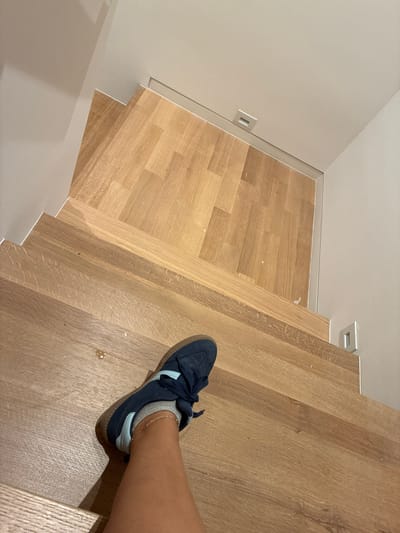 The image shows a person's leg and foot wearing a blue sneaker with white stripes and grey socks, standing on a wooden staircase. The staircase has a light brown, wood-grain finish and appears to be indoors. The wall to the left of the stairs is painted a neutral, light color and has a rectangular light fixture. The scene is well-lit, suggesting it is daytime or that there is artificial lighting. The focus is on the staircase and the person's footwear, with the background out of focus. There is no visible text in the image. The overall impression is of a clean, modern interior space. The image does not provide clues about the time of day, weather, or emotions.