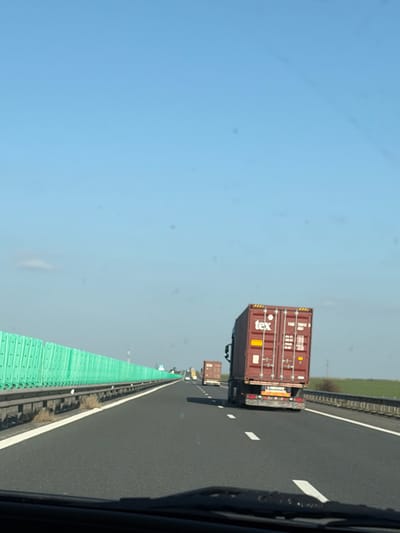 The image depicts a highway scene within the vicinity of Borcea, Romania, under clear daylight. The viewpoint is from inside a vehicle, looking forward. The sky is bright blue, containing several circular lens artifacts.
The highway is a multi-lane asphalt road. The lane immediately ahead is delineated by white dashed lines, and a solid white line borders the right shoulder. The lower portion of the image displays the dark top edge of a vehicle dashboard and a windshield wiper arm.
In the foreground, directly ahead in the right lane, is a semi-trailer truck hauling a red intermodal shipping container. The container's rear doors feature the white "tex" logo, identification numbers (e.g., "TCRU 200412 8B1"), and a vertical yellow stripe. The truck's taillights and mud flaps are visible. Further down the right lane, a second semi-trailer truck with a similar red container is present, followed by a third, less distinct vehicle in the far distance.
To the left of the road, a long, continuous green modular barrier, consistent with a sound or wind barrier, extends into the distance. Beyond the right shoulder, a strip of dry, light-colored vegetation transitions into an expanse of green fields. No individuals are visible. The scene indicates vehicular traffic in motion on the highway.