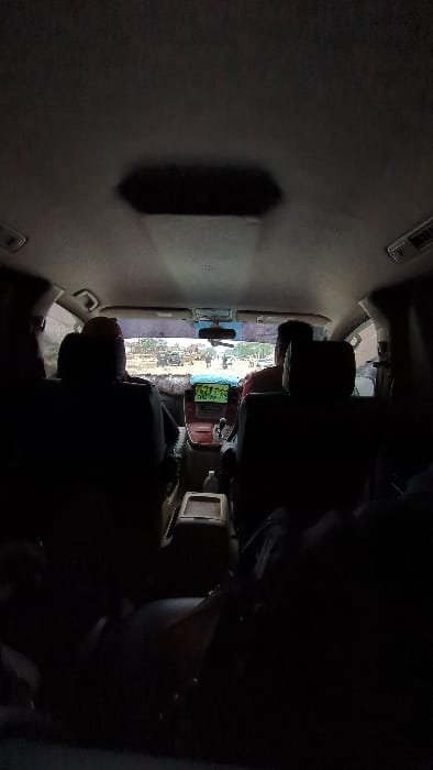 The image depicts the interior of a vehicle, likely a car, with a driver visible in the driver's seat. The dashboard features a screen displaying green text or graphics. Outside the windshield, there's a glimpse of what appears to be a construction site or outdoor area with vehicles and equipment. The interior is generally dark, and the lighting is poor, making it difficult to discern finer details. The seating arrangement suggests a typical passenger vehicle layout.