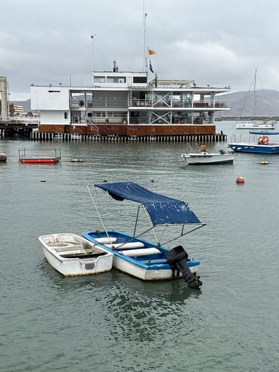 The image depicts a cloudy day at the port of La Punta, Peru. In the foreground, two small boats are moored in the water. The boat on the left is white and appears empty. The boat on the right is blue and white and has a blue canopy. It is equipped with a black outboard motor. In the midground, a large, multi-story white building with extensive glass windows stands on stilts over the water. A pier extends from the left side of the frame to this building. Several other boats and buoys are visible in the water in the midground and background, including a white sailboat with the registration "AYO." The background shows a hilly coastline under a gray, overcast sky.