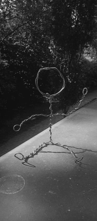 The black and white, medium-shot image shows a wire figure of a person sitting on a flat, textured surface. The figure is made of twisted wire, forming a circular head, a body, arms, and legs. The figure's arms are outstretched, with one arm bent upwards and the other extended to the side. The legs are bent at the knees, with the feet flat on the surface. The figure is casting a shadow on the surface, indicating it is outdoors and illuminated by sunlight.

The background is out of focus and appears to be foliage and trees, suggesting a natural setting. The overall mood of the image is somewhat somber due to the black and white tones and the solitary nature of the wire figure. The time of day is likely daytime, given the strong shadows. The location context of Karviná, Czechia, is provided, but no specific visual cues within the image directly identify this location. There is no visible text in the image.