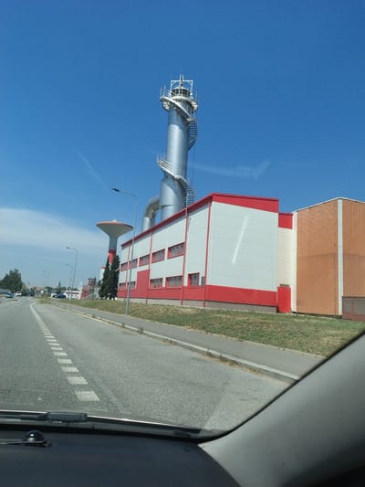 This media file captures an industrial scene under a clear, bright blue sky, suggesting a sunny day. The perspective is from inside a vehicle, indicated by the dark gray car interior elements on the right side of the frame and the windshield wipers. The main subjects are industrial buildings and a tall, cylindrical structure with a spiraling staircase.
One prominent building is a large, multi-story structure with a white facade accented by red trim. It features a series of rectangular windows with red frames. Adjacent to this is another building with a roof made of corrugated, reddish-brown material. To the left of the white and red building stands a tall, metallic industrial chimney or tower. This tower has a wide base that tapers upwards, and a prominent spiral staircase winds around its upper section, leading to a platform. Further down the road, a conical structure, possibly a water tower or part of the industrial complex, is visible.
The setting appears to be an industrial area bordering a paved road. The road has visible lane markings, including dashed white lines. On the side of the road, there's a strip of green grass with some sparse vegetation. In the distance, more buildings and trees can be seen, suggesting a town or city nearby. The overall atmosphere is functional and industrial, bathed in the bright light of daytime. No people are explicitly visible, and there is no discernible text in the image.