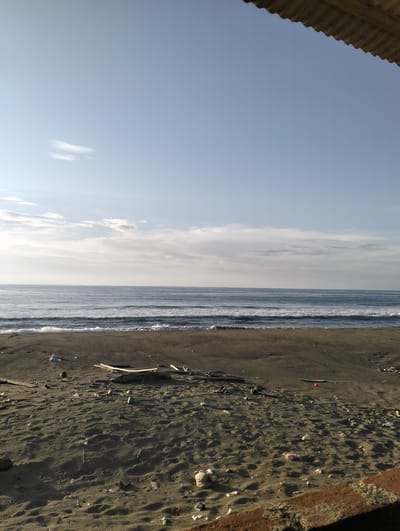 The image depicts a beach scene in Peuneulet Baroh, Indonesia. The sky is predominantly blue with scattered white clouds. Below the sky is a body of water with gentle waves breaking on the shore. The beach is covered in dark sand, with visible footprints and scattered debris including plastic waste, twigs, and what appears to be broken wood. A rusted corrugated metal structure is visible in the top right corner of the image and another rusted surface appears at the bottom right corner. There are no people present in the image. The scene appears to be a somewhat polluted beach environment.